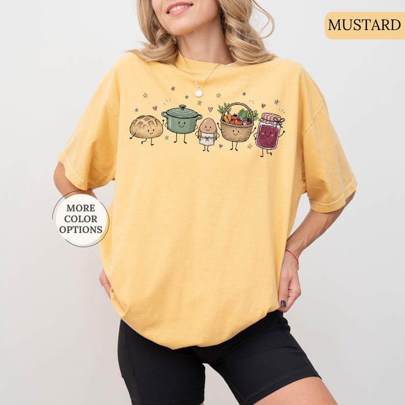 May include: A mustard yellow t-shirt featuring a graphic print of five anthropomorphic food items including a loaf of bread, a green pot, an egg, a basket of vegetables, and a jar of jam, all with small smiling faces, arms, and legs.