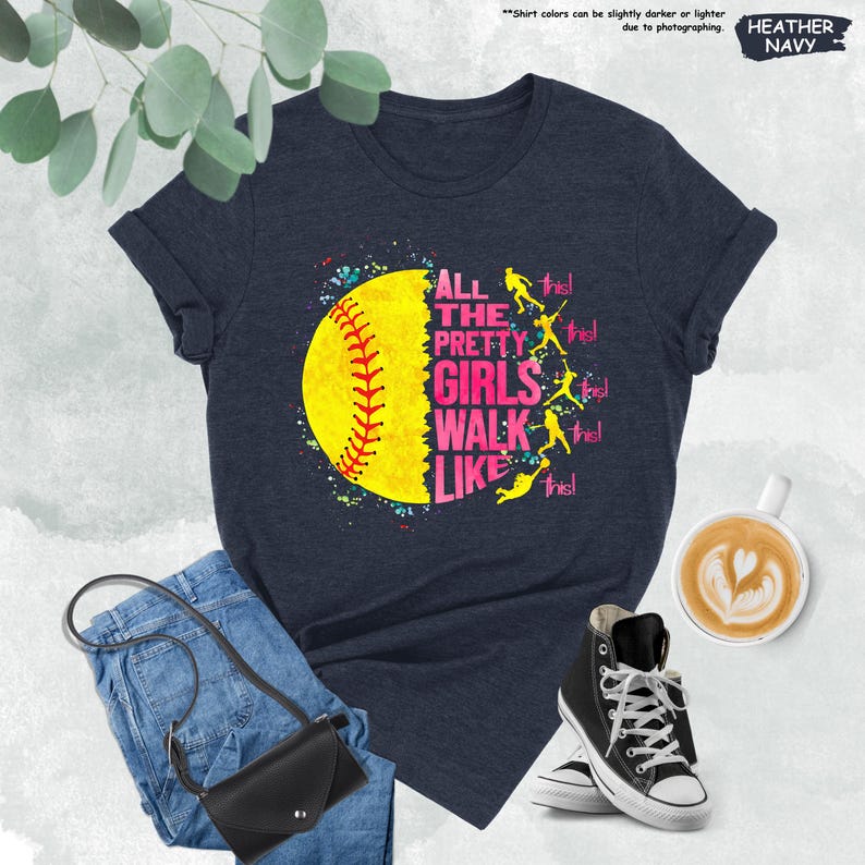 May include: Heather navy t-shirt with a softball graphic. The graphic features a yellow softball with red stitching and the text "ALL THE PRETTY GIRLS WALK LIKE THIS!" in pink and white. The shirt is paired with blue jeans, a black purse, and black high-top sneakers.