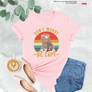 May include: Pink t-shirt with the text "DON'T WORRY - BE CAPY-" and a graphic of a capybara wearing sunglasses. The design is set against a retro sunset background. The shirt is paired with blue jeans and beige shoes.