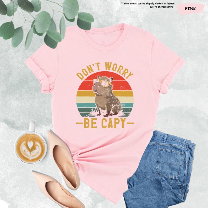 May include: Pink t-shirt with the text "DON'T WORRY - BE CAPY-" and a graphic of a capybara wearing sunglasses. The design is set against a retro sunset background. The shirt is paired with blue jeans and beige shoes.