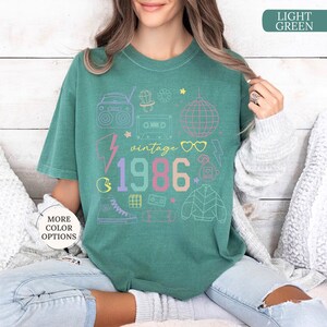May include: A light green t-shirt with a vintage 1986 graphic design. The design includes a boombox, cassette tape, disco ball, and other 80s-themed illustrations in pastel colors. The shirt is a relaxed fit.