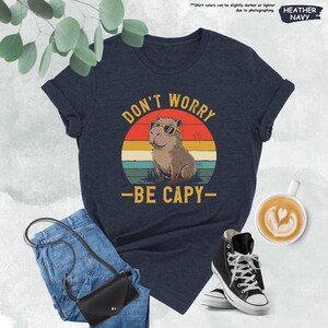 May include: Heather navy t-shirt with a graphic of a capybara wearing sunglasses, set against a sunset design. The text reads "DON'T WORRY - BE CAPY-". The shirt is paired with jeans, a black purse, and black high-top sneakers.