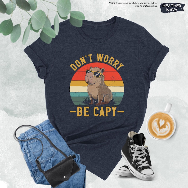 May include: Heather navy t-shirt with a graphic of a capybara wearing sunglasses, set against a sunset design. The text reads "DON'T WORRY - BE CAPY-". The shirt is paired with jeans, a black purse, and black high-top sneakers.