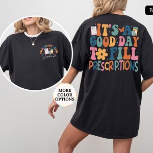 May include: Black t-shirt with the text "IT'S A GOOD DAY TO FILL PRESCRIPTIONS" in colourful, retro-style lettering on the back. The front features a smaller design with "PHARMACY TECH" and a name. A fun, casual tee.