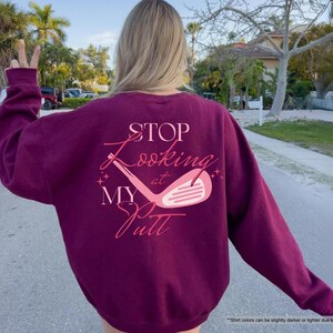 以下が含まれることがあります： ピンク色のスクリプトで「STOP Looking at MY Putt」というテキストとゴルフクラブのグラフィックが描かれた、マルーン色のスウェットシャツ。スウェットシャツは無地で、長袖です。