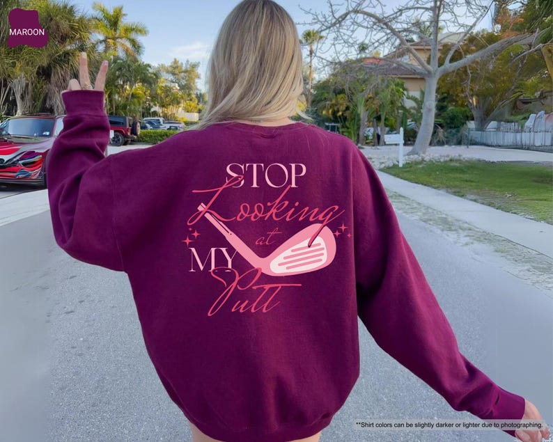 以下が含まれることがあります： ピンク色のスクリプトで「STOP Looking at MY Putt」というテキストとゴルフクラブのグラフィックが描かれた、マルーン色のスウェットシャツ。スウェットシャツは無地で、長袖です。