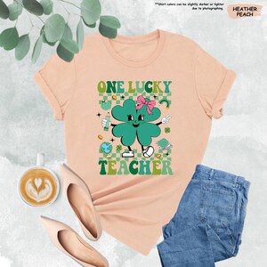 May include: A peach colored t-shirt with a green four-leaf clover cartoon character wearing a pink bow. The clover is holding a green shamrock and a peace sign. The text "ONE LUCKY TEACHER" is printed on the shirt.