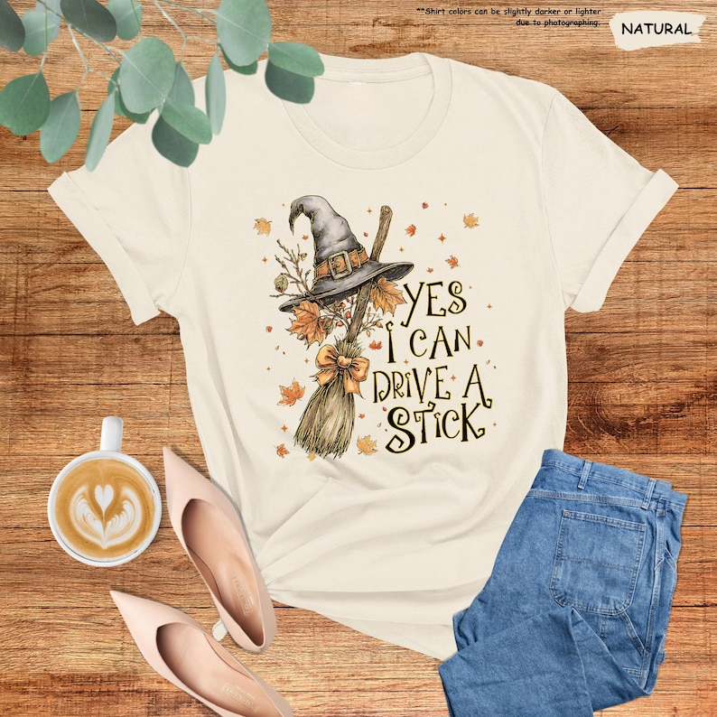 May include: A natural-colored t-shirt featuring a graphic of a witch hat resting on a broomstick with autumn leaves and the text "YES I CAN DRIVE A STICK" in a dark font. The design includes a small orange bow tied around the broom handle.