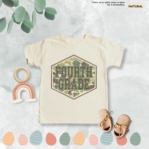 May include: A cream-coloured Fourth Grade t-shirt featuring a camouflage design within a hexagonal frame. The text "FOURTH GRADE" is in a green, distressed font. The image includes a wooden rainbow, baby shoes, and decorative Easter eggs.