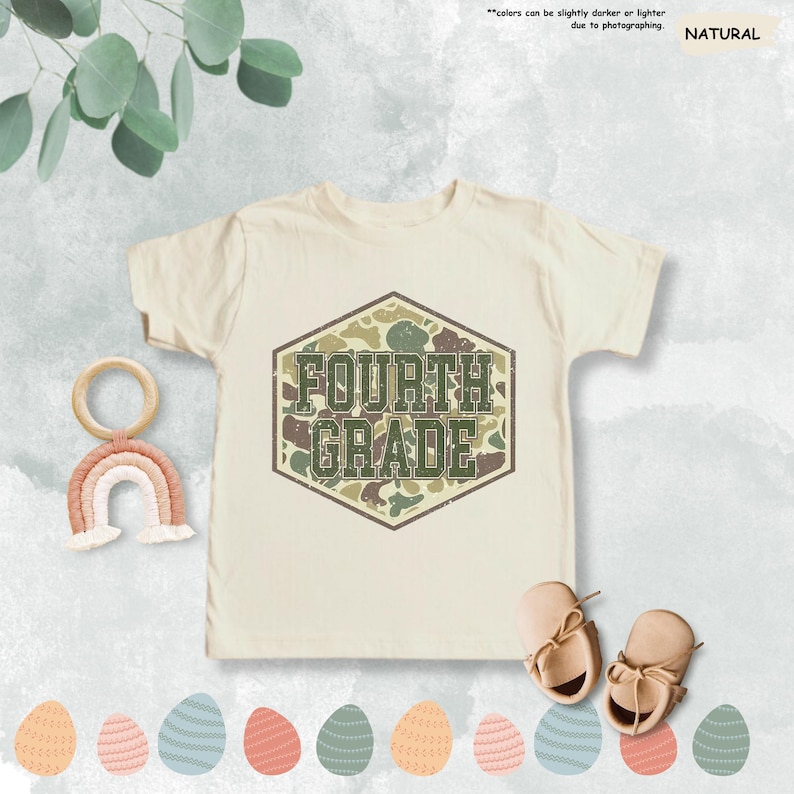 May include: A cream-coloured Fourth Grade t-shirt featuring a camouflage design within a hexagonal frame. The text "FOURTH GRADE" is in a green, distressed font. The image includes a wooden rainbow, baby shoes, and decorative Easter eggs.
