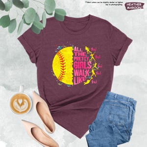 May include: Maroon t-shirt with the text "ALL THE PRETTY GIRLS WALK LIKE THIS!" and a yellow softball graphic. The shirt is paired with blue jeans and nude-colored flats. The words "HEATHER MAROON" are also visible.