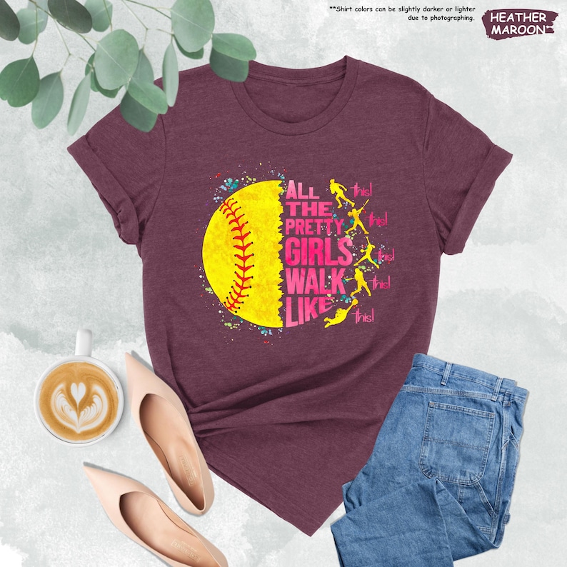 May include: Maroon t-shirt with the text "ALL THE PRETTY GIRLS WALK LIKE THIS!" and a yellow softball graphic. The shirt is paired with blue jeans and nude-colored flats. The words "HEATHER MAROON" are also visible.