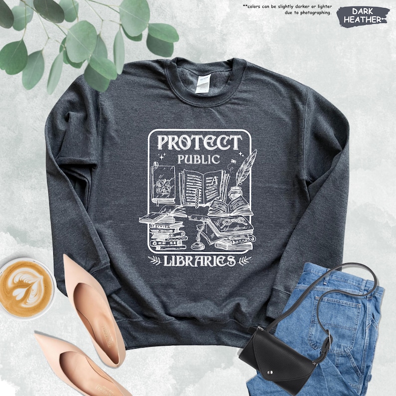 May include: Dark heather grey sweatshirt with a white graphic design featuring a stack of books, a quill pen, and the text "Protect Public Libraries".