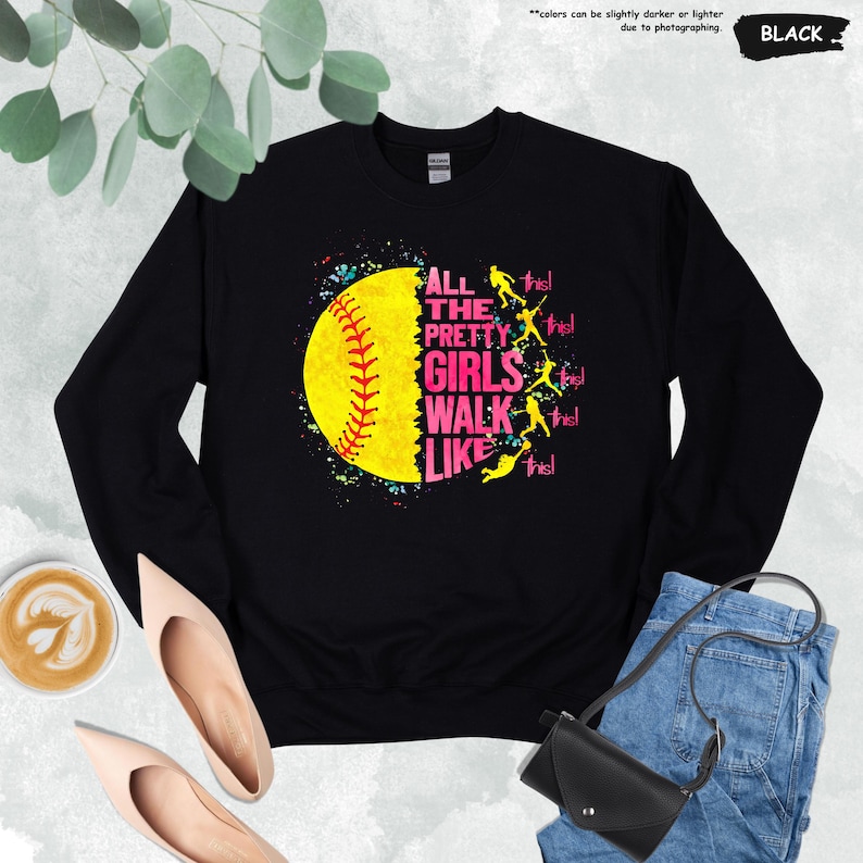 May include: Black sweatshirt featuring a large yellow softball graphic with red stitching and the text "ALL THE PRETTY GIRLS WALK LIKE THIS!" in pink. The design includes colorful paint splatters and silhouettes of softball players.