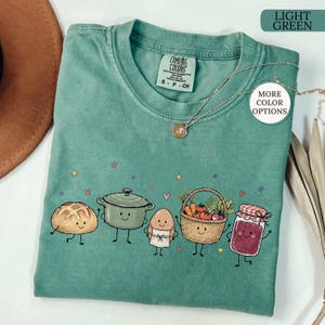 May include: A light green crew neck t-shirt featuring a graphic print of a smiling loaf of bread, a cooking pot, an egg in an apron, a basket of vegetables, and a jar of jam. The shirt includes a tag reading Comfort Colors and a gold necklace.