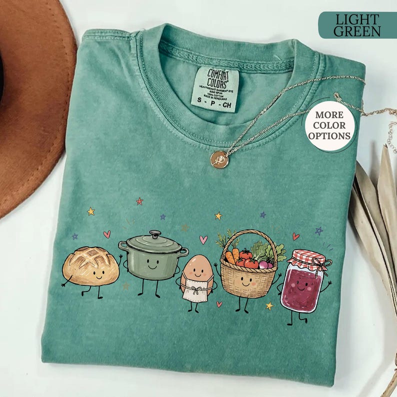 May include: A light green crew neck t-shirt featuring a graphic print of a smiling loaf of bread, a cooking pot, an egg in an apron, a basket of vegetables, and a jar of jam. The shirt includes a tag reading Comfort Colors and a gold necklace.