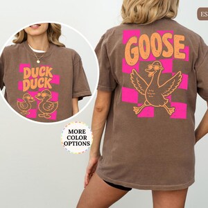 May include: An espresso brown t-shirt featuring a back graphic of a goose wearing sunglasses over a pink checkered pattern with the text GOOSE. The front chest graphic shows two ducks wearing sunglasses with the text DUCK DUCK.