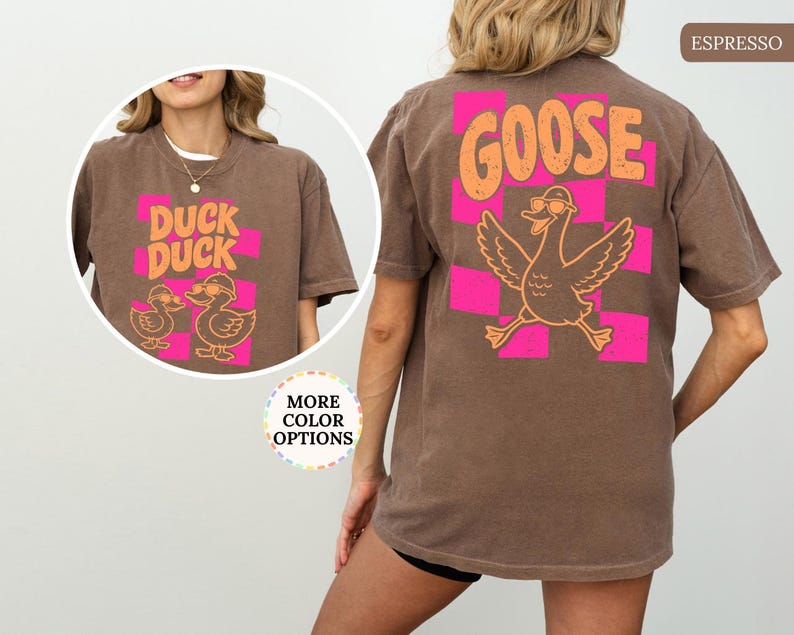 May include: An espresso brown t-shirt featuring a back graphic of a goose wearing sunglasses over a pink checkered pattern with the text GOOSE. The front chest graphic shows two ducks wearing sunglasses with the text DUCK DUCK.