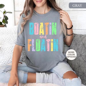 May include: A grey t-shirt featuring the text "BOATIN and FLOATIN" in a distressed, multi-coloured font. The short-sleeved shirt has a relaxed fit. The text uses shades of blue, yellow, pink, and green.