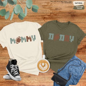 May include: Two t-shirts, one white and one green, with the words "MOMMY" and "DDDY" printed on them. The word "MOMMY" has a football graphic next to it. The word "DDDY" has a football graphic in the middle of the word.