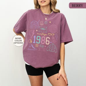 May include: Berry-colored vintage-themed t-shirt with a white undershirt. The shirt features a colorful graphic with the text "vintage 1986" and various 80s-themed illustrations, including a boombox, cassette tape, and disco ball.