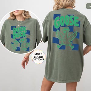 May include: A moss green short-sleeve t-shirt featuring a blue checkered pattern on the back with a green graphic of a goose and the text GOOSE. The front includes a circular inset showing the same shirt with a duck graphic and the text DUCK DUCK.