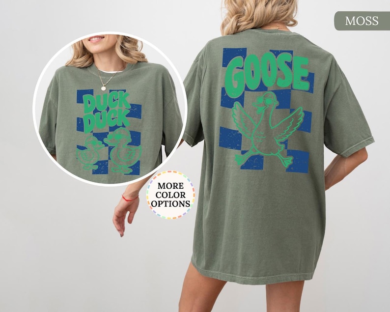 May include: A moss green short-sleeve t-shirt featuring a blue checkered pattern on the back with a green graphic of a goose and the text GOOSE. The front includes a circular inset showing the same shirt with a duck graphic and the text DUCK DUCK.