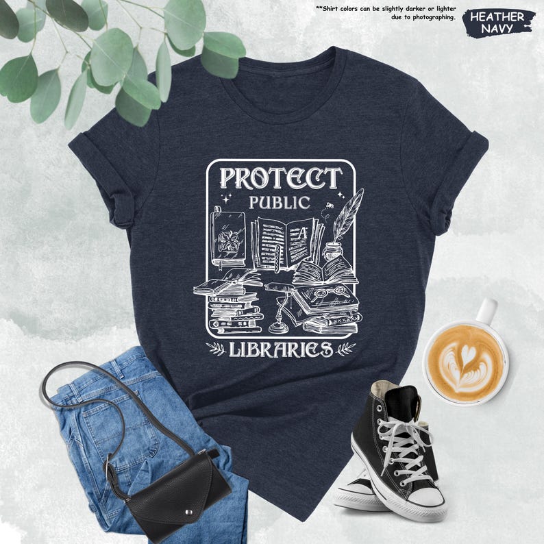 May include: A heather navy blue t-shirt with a white graphic design of a stack of books, a quill pen, and a potion bottle. The graphic also includes the text "Protect Public Libraries".