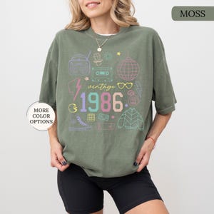 May include: Moss green t-shirt with a colorful retro graphic. The design includes a boombox, cassette tape, disco ball, and the year "1986." Additional text reads "vintage." The shirt has a relaxed fit and short sleeves. "More color options" text is visible.