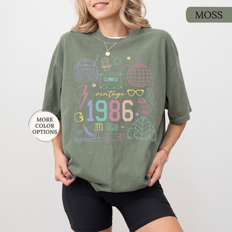 May include: Moss green t-shirt with a colorful retro graphic. The design includes a boombox, cassette tape, disco ball, and the year "1986." Additional text reads "vintage." The shirt has a relaxed fit and short sleeves. "More color options" text is visible.