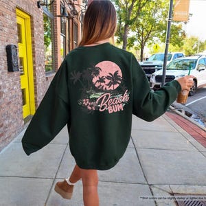May include: Dark green sweatshirt featuring a beach-themed design in pink and white on the back. The graphic includes palm trees, a sunset, and the words "Beach Bum". The sweatshirt is a casual style.
