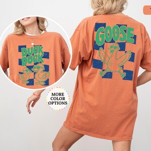 May include: An orange t-shirt featuring a graphic on the back and front. The back displays the word GOOSE in green above a green goose wearing sunglasses over a blue checkered pattern. The front shows DUCK DUCK with two ducks in sunglasses.