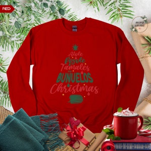 May include: Red long-sleeved sweatshirt with a green and pink Christmas tree design. The tree is made up of the words "Atole", "Pozole", "Tamales", "Bu&ntilde;uelos", and "Christmas".