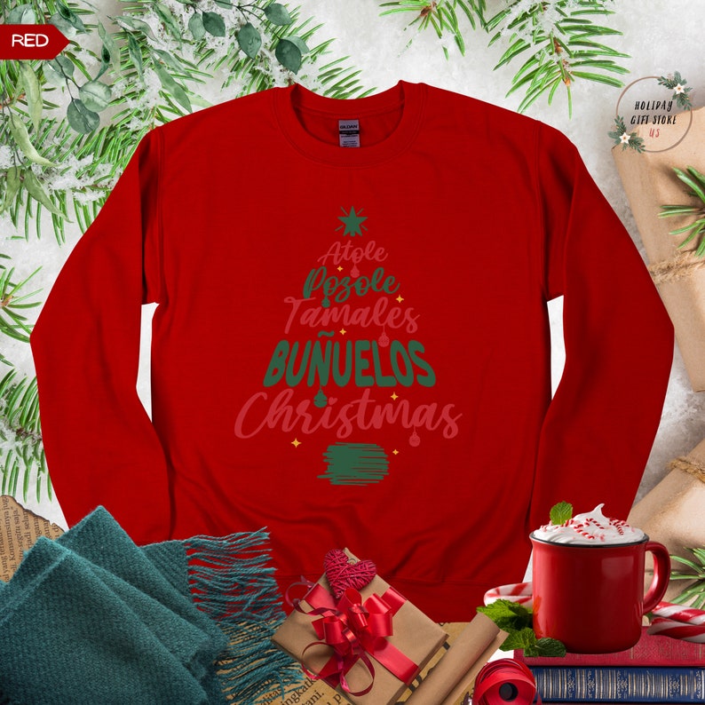 May include: Red long-sleeved sweatshirt with a green and pink Christmas tree design. The tree is made up of the words "Atole", "Pozole", "Tamales", "Bu&ntilde;uelos", and "Christmas".