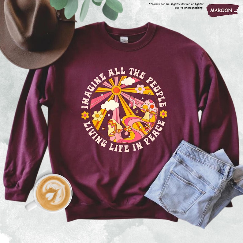 May include: A maroon sweatshirt with a retro-style graphic design featuring a sun, clouds, mushrooms, and flowers. The text "Imagine All The People Living Life In Peace" is printed in a circle around the graphic.