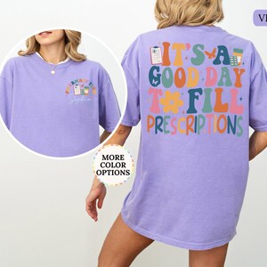 May include: A violet t-shirt with the text "IT'S A GOOD DAY TO FILL PRESCRIPTIONS" in colourful, retro-style lettering on the back. The front features a smaller design with pharmacy-related graphics. The shirt has a comfortable, oversized fit.