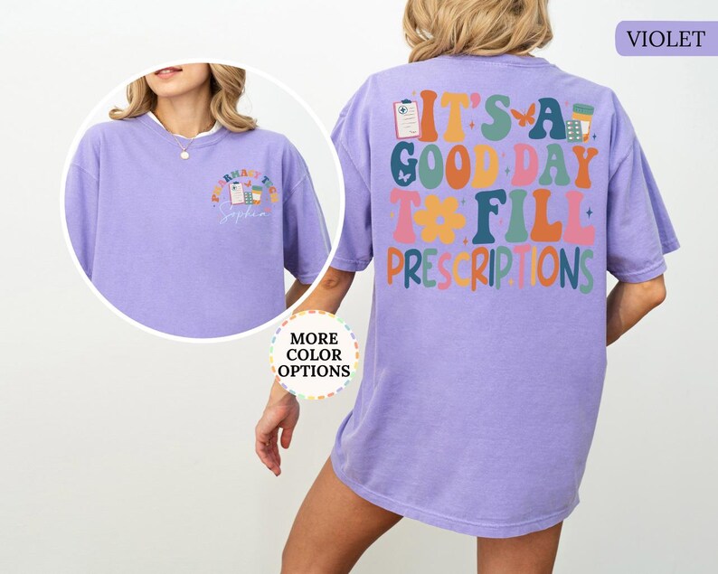 May include: A violet t-shirt with the text "IT'S A GOOD DAY TO FILL PRESCRIPTIONS" in colourful, retro-style lettering on the back. The front features a smaller design with pharmacy-related graphics. The shirt has a comfortable, oversized fit.