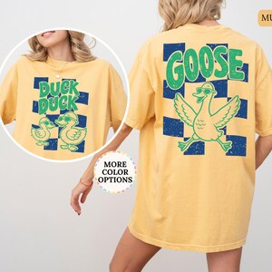 May include: A mustard yellow t-shirt featuring a dark blue checkered pattern on the back with a large green goose graphic and the text GOOSE. The front inset shows the same shirt with two ducks and the text DUCK DUCK in green.
