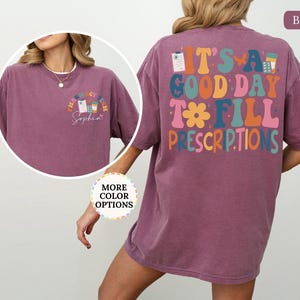 May include: Berry-coloured t-shirt with the text "It's a good day to fill prescriptions" on the back in colourful, retro-style lettering. The front features a smaller design with pharmacy-related graphics and the name "Sophia".