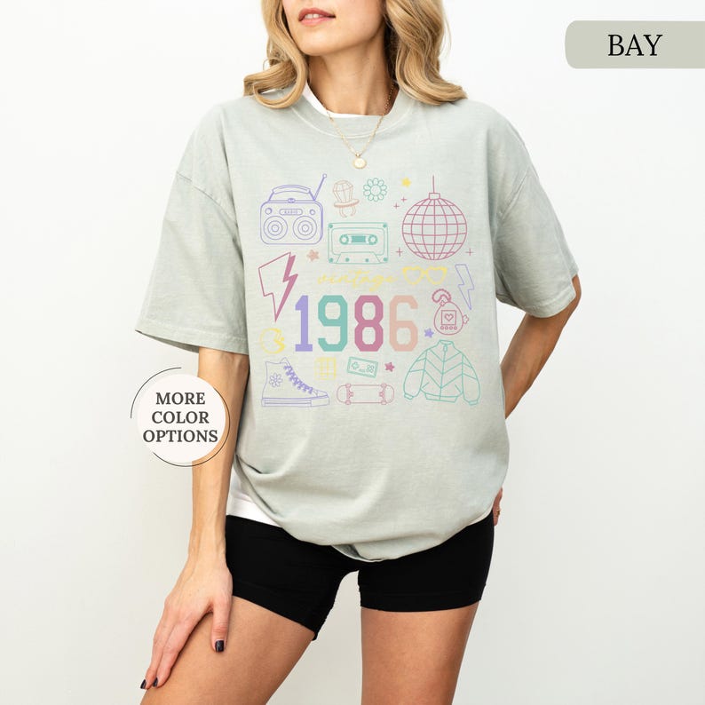 May include: A light green t-shirt with a retro 1986 graphic. The design includes a boombox, cassette tape, disco ball, and other 80s-themed icons in pastel colors. The shirt is a relaxed fit.