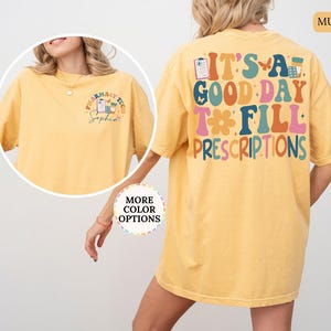 May include: Mustard-coloured t-shirt with the text "IT'S A GOOD DAY TO FILL PRESCRIPTIONS" in colourful, retro-style lettering. The front of the shirt features a pharmacy tech design. The shirt is a comfortable, oversized fit.