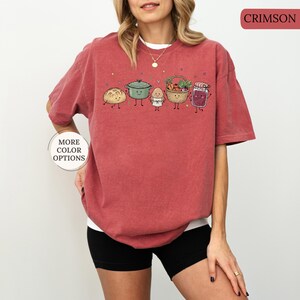 May include: A crimson short-sleeve t-shirt featuring a graphic print of five anthropomorphic food items including a loaf of bread, a green pot, an egg, a basket of vegetables, and a jar of jam. The text "CRIMSON" and "MORE COLOR OPTIONS" is visible.
