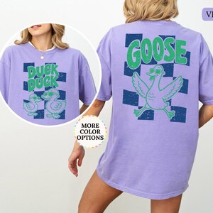 May include: A violet t-shirt featuring a graphic on the back with a large green goose wearing sunglasses over a dark blue checkered pattern and the text GOOSE. A circular inset shows the front of the shirt with two ducks and the text DUCK DUCK.