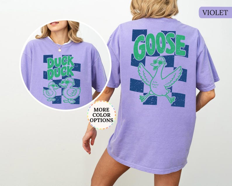 May include: A violet t-shirt featuring a graphic on the back with a large green goose wearing sunglasses over a dark blue checkered pattern and the text GOOSE. A circular inset shows the front of the shirt with two ducks and the text DUCK DUCK.