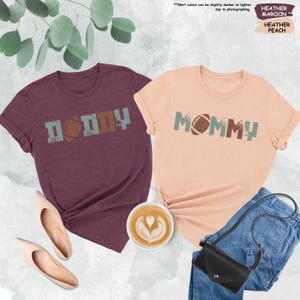 May include: Two t-shirts with a football theme. The maroon shirt says "DADDY" with a football in the middle. The peach shirt says "MOMMY" with a football in the middle.