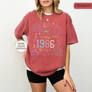 May include: Crimson-colored t-shirt with a vintage 1986 graphic. The design includes a boombox, cassette tape, disco ball, and other retro icons. The shirt is a relaxed fit and has short sleeves. The words "More Color Options" are also visible.