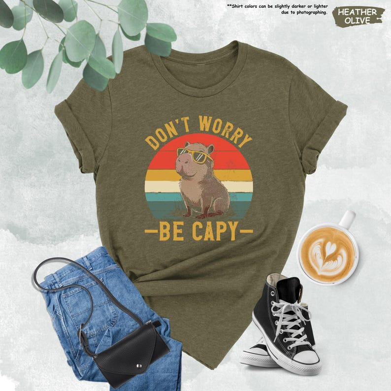 May include: Heather olive green t-shirt with a graphic of a capybara wearing sunglasses, set against a retro sunset design. The text reads "DON'T WORRY - BE CAPY-". The shirt is paired with blue jeans, a black purse, and black high-top sneakers.