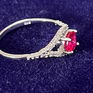 May include: A silver ring with a vibrant red oval gemstone. The band has a delicate, textured design with small, sparkling accents. The ring is set against a dark blue background.