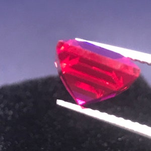 May include: A heart-shaped, faceted gemstone in a vivid red colour, held by tweezers. The gem's facets reflect light, creating a play of colour. The background is a dark, blurred texture.