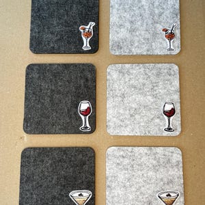 Puede incluir: Seis posavasos cuadrados, tres grises oscuros y tres grises claros, están dispuestos en dos filas. Cada posavasos presenta una pegatina de copa de cóctel. Las pegatinas representan un cóctel, una copa de vino tinto y una copa de martini.